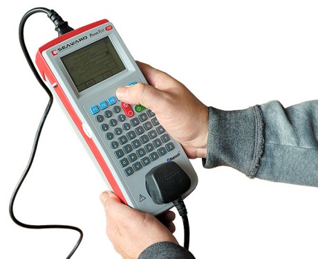 A hand holding a portable appliance tester, with a 13 amp plug held in its test socket